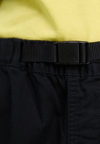 Schwarze Shorts mit einem strukturierten Bund und einer Kunststoffschnalle, vor einem gelben Hemd-Hintergrund, der ein einfaches Design zeigt.