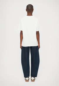 T-shirt bianco a maniche corte indossato con jeans larghi di colore blu scuro. Le calzature presentano una combinazione di accenti bianchi e verde oliva.