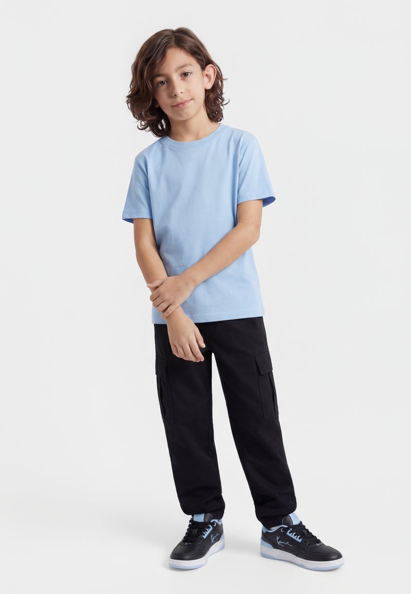 Chlapec se středně dlouhými hnědými vlasy, který má na sobě světle modré tričko, černé kalhoty s kapsami a černé tenisky s modrými detaily, stojící na bílém pozadí.