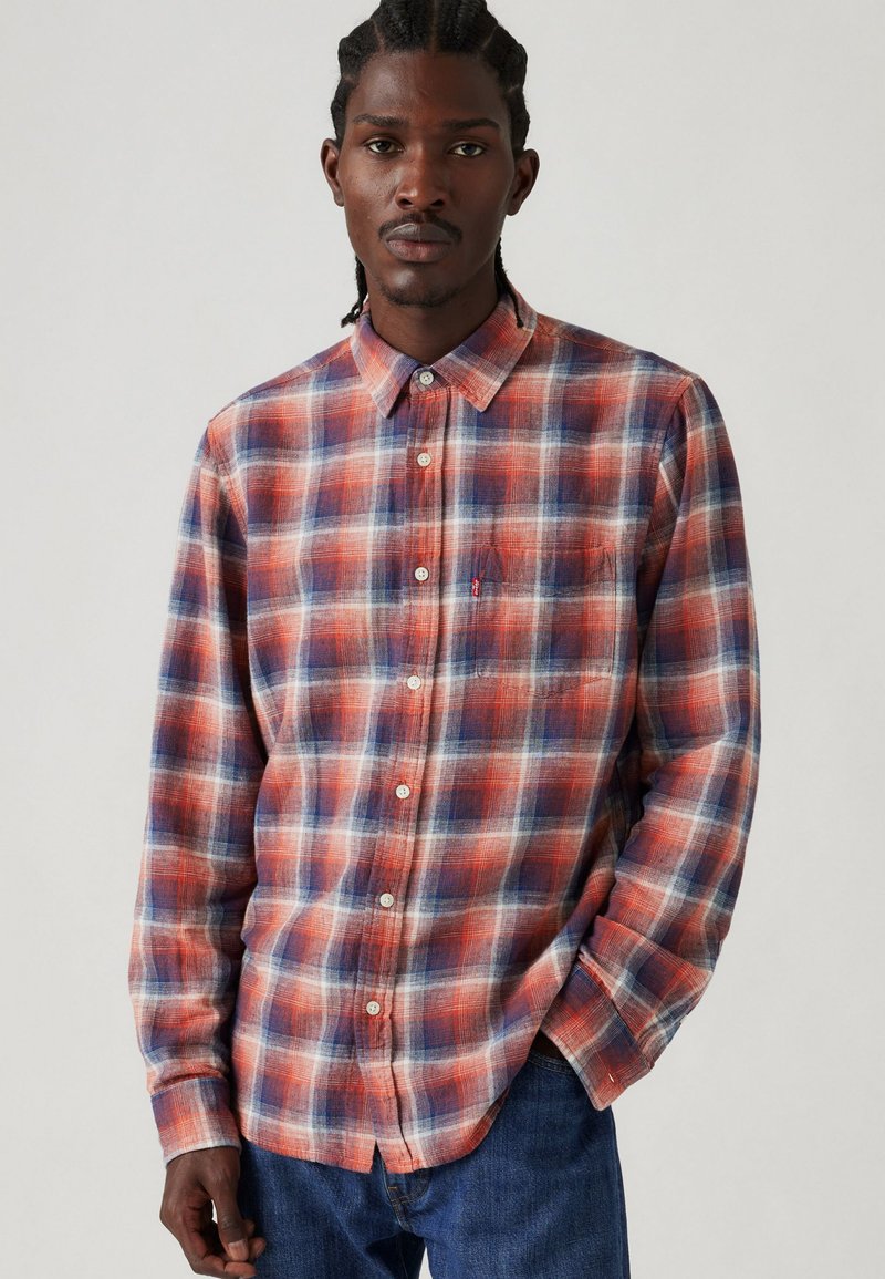 Chemise à boutons à carreaux en rouge, bleu et blanc ; fabriquée en tissu léger avec un col et des manches longues. Comprend un détail de poche à l'avant.