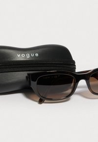 Čierne slnečné okuliare s elegantným, obdĺžnikovým rámom a tónovanými sklami, ležiace vedľa čierneho zipsového puzdra označeného "vogue."