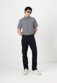 Camicia a maniche corte con bottoni, con motivo a foglie navy, abbinata a pantaloni scuri e sneakers bianche. Il modello indossa occhiali da sole.