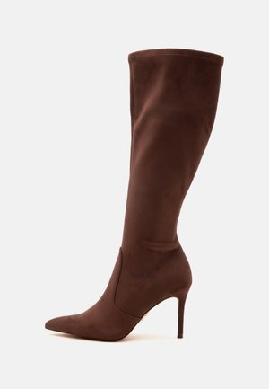 Botte marron pour femme montant jusqu’au genou avec bout pointu et talon haut et fin sur un fond uni.