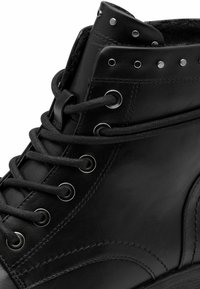 Botas de cuero negras hasta el tobillo con cordones redondos, ojales plateados y tachuelas decorativas cerca de la parte superior. Textura suave con detalle de costura curvada.