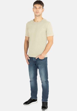 Beige Baumwoll-T-Shirt mit Rundhals, kurzen Ärmeln, kombiniert mit schmal geschnittenen blauen Jeans und schwarzen Sneakers mit weißen Sohlen.