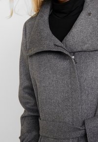 Cappotto grigio in misto lana con un grande colletto a risvolto, chiusura con zip e vita con cintura. Tessuto testurizzato con un sottile motivo a zig zag.