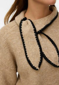 Beżowy sweter z dzianiny z dużym kołnierzem i czarną falbanką, zawiązywany pod szyją. Miękka faktura i luźny krój.