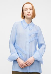 Νέος/νέα με κοντά κόκκινα μαλλιά, φοράει διαφανές ανοιχτό γαλάζιο πουκάμισο με κρόσσια στο γιακά, τις μανσέτες και το μπροστινό μέρος, με τα χέρια ενωμένα.