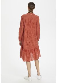 Šaty v rustikální barvě s dlouhými rukávy, krajkovou tkaninou, nařaseným živůtkem a volánkovým lemem, doplněné bílými teniskami.