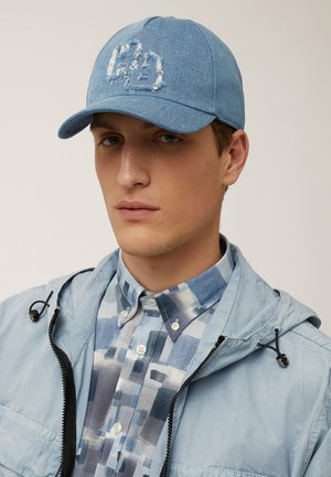 Light blue denim cap with a distressed embroidered logo. Features a curved visor and textured fabric, paired with a patterned button-up shirt.