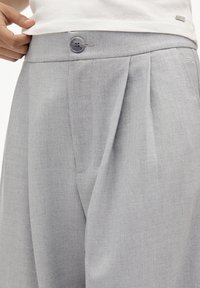 Pantalon gris clair ajusté avec une fermeture à un bouton et des plis à l'avant, confectionné dans un tissu lisse et texturé.