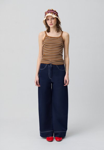 Débardeur brun rayé, pantalon large navy avec surpiqûres blanches, et chaussures rouges ; le modèle porte un chapeau de seau décoratif beige.