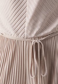 Beige dress featuring vertical ribbed texture, fitted bodice, and a tie belt at the waist, with flowing pleats starting from the waist.