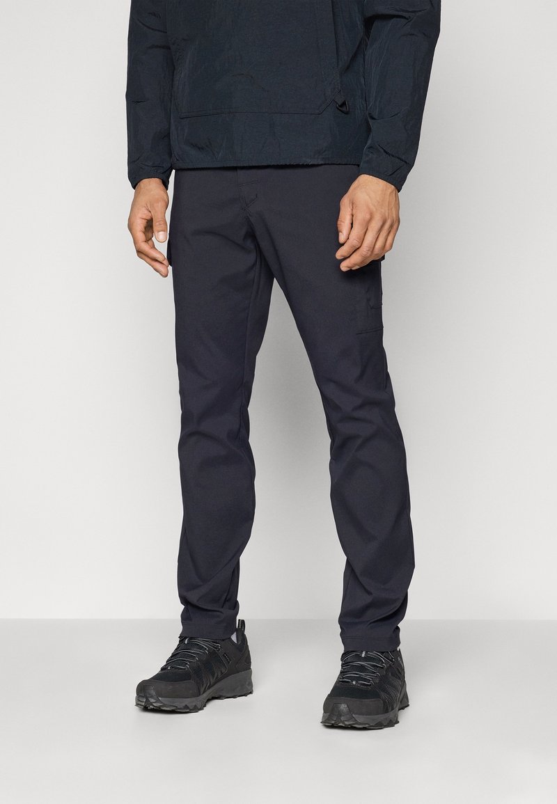 Man med svarta cargobyxor, svart långärmad jacka och svarta träningsskor står framför en neutral bakgrund.