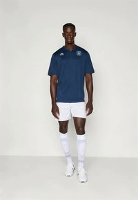 Tamsiai mėlyna polo marškinėliai su apykakle, derinami su baltais šortais ir raštuotomis baltomis kojinėmis. Apsirengta baltais sportiniais bateliais.