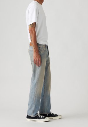 Mann steht seitlich, trägt ein weißes T-Shirt, hellblaue ausgewaschene Jeans, schwarze Sneakers mit niedrigem Schnitt, sichtbare Tattoos am linken Arm.