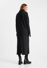 Zwart gebreide jurk met hoge kraag, lange mouwen en zijsplitten. Beschikt over een ribbelzoom en is gecombineerd met zwarte enkellaarsjes.