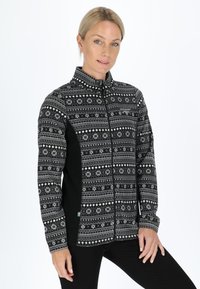 Woman wearing a black and white patterned zip-up fleece jacket with snowflake designs, standing with one hand on her hip against a white background.