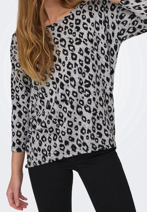 Femme portant un haut gris à manches longues avec des taches léopard noires et un pantalon noir, posant avec un bras levé.