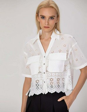 Femme blonde portant une chemise blanche à manches courtes avec motifs floraux ajourés et deux poches poitrine, associée à un pantalon noir.
