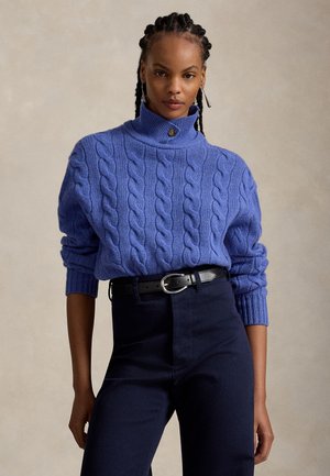 Frau mit geflochtenem Haar, die einen blauen Zopfmuster-Rollkragenpullover, einen schwarzen Gürtel und dunkle hoch taillierte Hosen vor einem einfarbigen Hintergrund trägt.