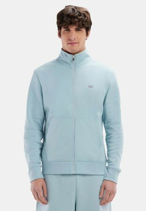 Sweat-shirt zippé bleu clair en tissu doux avec un col haut, des poignets côtelés et un petit logo sur la poitrine. Il est doté de poches latérales.