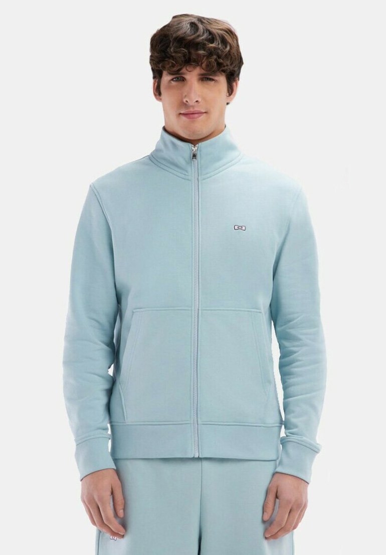 Sweat-shirt zippé bleu clair en tissu doux avec un col haut, des poignets côtelés et un petit logo sur la poitrine. Il est doté de poches latérales.
