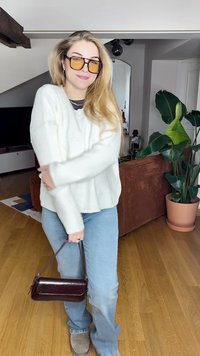 Biała sweter na prążki, jasnoniebieskie dżinsy, szare obuwie i mała bordowa torebka. Akcesoria to dużej wielkości żółte okulary przeciwsłoneczne. Wnętrze.