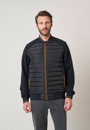 Man met baard die een zwart doorgestikt jack met oranje ritssluitdetails en donkergrijze broek draagt tegen een effen lichte achtergrond.