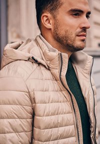 Beige pufferjack met een ritssluiting aan de voorkant en een capuchon, met horizontale stitching, gedragen over een donkergroene geribde trui.