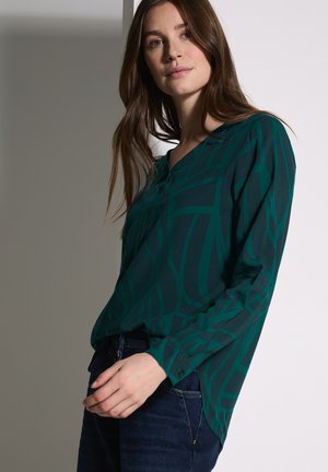 Blusa teal con un motivo geometrico, maniche lunghe e scollatura a V, realizzata in tessuto morbido, abbinata a jeans in denim scuri.