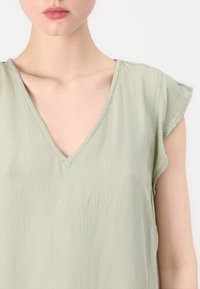 Lys grønn, teksturert bluse med V-hals og korte flatterende ermer. Stoffet ser mykt og litt krøllete ut, noe som gir visuell interesse.
