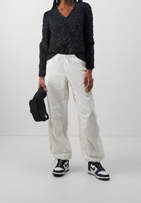 Top lungo a maniche lunghe nero con texture, pantaloni bianchi larghi e sneakers nere con dettagli bianchi. Modella che tiene una piccola borsa nera.