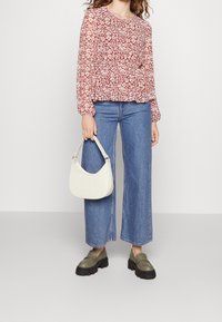 Donna che indossa una blusa lunga a maniche lunghe rossa a fiori, jeans larghi blu, mocassini olive con zeppa, mentre tiene una borsa bianca su uno sfondo semplice.