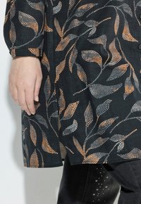 Main gauche reposant à côté d'un manteau noir avec des motifs de feuilles tachetés d'orange et de gris, associé à un pantalon noir doté de petits points blancs.