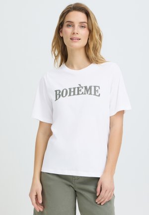 Giovane donna con capelli biondi che indossa una T-shirt bianca con la scritta "BOHÈME" e pantaloni verde oliva, in piedi davanti a uno sfondo chiaro uniforme.
