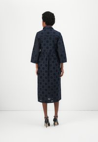 Abito midi blu navy con motivo floreale a occhiello, maniche a tre quarti e vita arricciata. Indossato con scarpe con tacco alto nere.