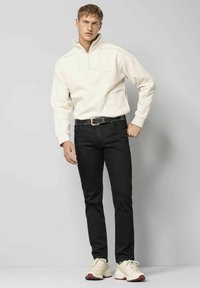 Sweat à fermeture éclair beige clair avec poches latérales, associé à un jean noir slim et des baskets crème avec des accents rouges, sur un fond neutre.