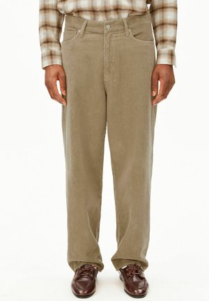Mann trägt beige Cordhose, braune Lederschuhe und ein beige-braunes kariertes Langarmhemd mit entspannten Händen an den Seiten.