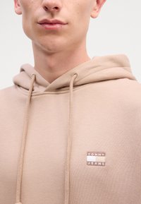 Hanorac bej cu șnur. Dispune de o aplicație rectangulară maro cu accente albe și bej, inscripționată "TOMMY JEANS." Textură netedă.