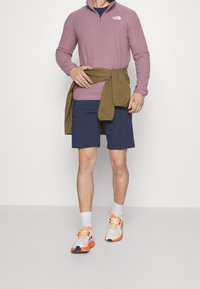 Langærmet, lilla zip-up skjorte, marineblå shorts, olivengrøn sweatshirt bundet om taljen, hvide sneakers med orange detaljer. Enkelt baggrund.