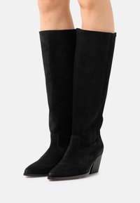 Bottes noir en suède montantes avec un bout pointu et un talon bloc modéré. Texture lisse avec un design épuré et minimaliste.