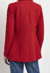 Personne portant un blazer rouge texturé et un jean bleu, vue de dos sur un fond blanc uni.