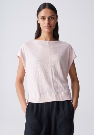 Femme aux longs cheveux foncés portant un haut rose pâle à manches courtes et un pantalon noir, debout les mains dans les poches devant un fond uni.