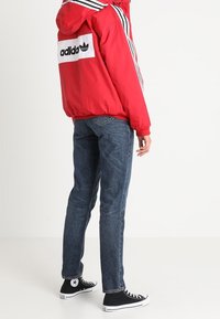 Rød hættejakke med hvidt logo og sorte striber, blå jeans og sorte lavprofil sneakers med hvide detaljer. Sidevinkel.