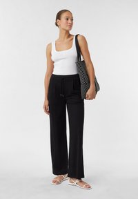 Black wide-leg pants paired with a white fitted tank top. The model holds a black and white striped tote bag and wears white strappy sandals.