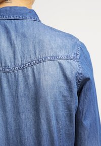 Camicia di jeans azzurro chiaro con colletto classico, cuciture visibili e tessuto testurizzato, che mostra la vista posteriore e parte della spalla.