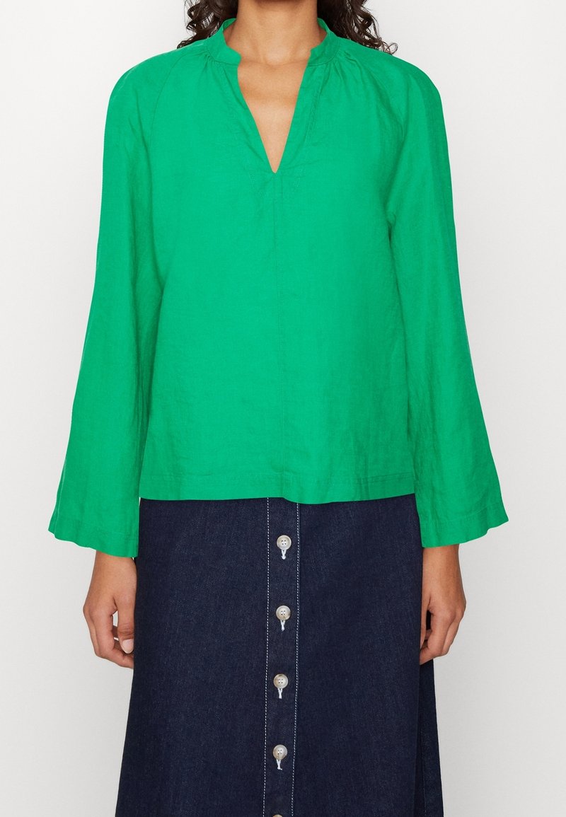 Blusa verde de lino con escote en V y detalles fruncidos en el escote. Mangas largas acampanadas y un corte holgado, combinada con una falda vaquera.