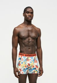 Μυώδης άνδρας στέκεται μπροστά σε λευκό φόντο, φοράει πολύχρωμα μπόξερ με σχέδια και πορτοκαλί μέση Björn Borg.