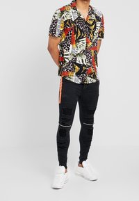 Camisa de manga corta multicolor con patrones geométricos, combinada con jeans negros desgastados que tienen una franja lateral naranja y zapatillas blancas.